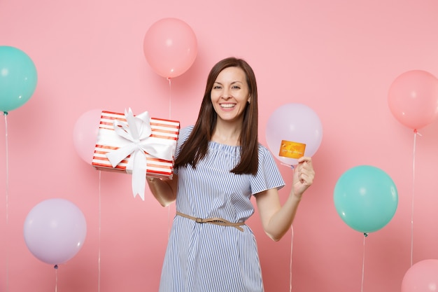 Portret van mooie lachende jonge vrouw in blauwe jurk met creditcard en rode doos met cadeau aanwezig op roze achtergrond met kleurrijke luchtballon. Verjaardagsfeestje, oprechte emoties van mensen.