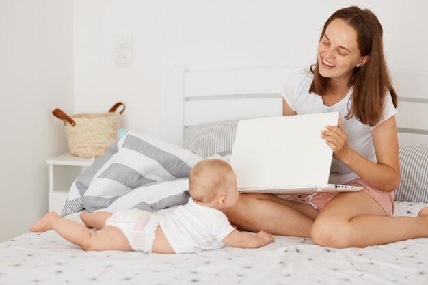 Portret van mooie lachende vrolijke vrouw met kleine neaborn baby zittend op het bed met laptop, jonge volwassen moeder probeert te werken en tijd door te brengen met dochtertje.