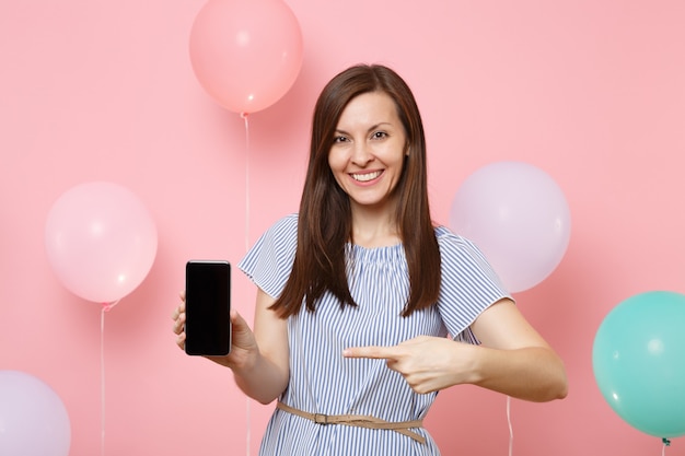 Portret van mooie lachende vrouw in blauwe jurk met wijzende wijsvinger op mobiele telefoon met leeg leeg scherm op roze achtergrond met kleurrijke luchtballonnen. verjaardagsfeestje concept.