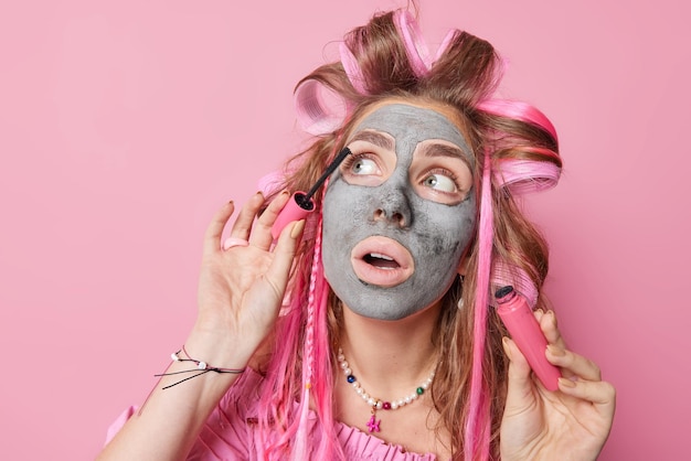 Portret van mooie langharige jonge vrouw past krulspelden toe voor het maken van kapsel mascara voert dagelijkse ochtendroutine zet op cosmetisch product ziet er verbaasd weg geïsoleerd over roze achtergrond