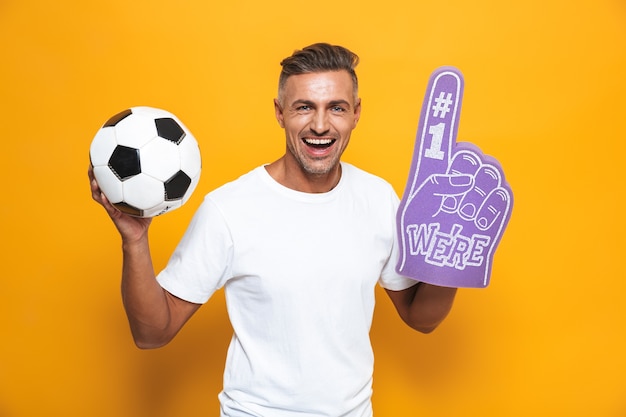 Foto portret van mooie man 30s in wit t-shirt met voetbal en nummer één fan hand handschoen met vinger aan de orde gesteld terwijl staande geïsoleerd op geel