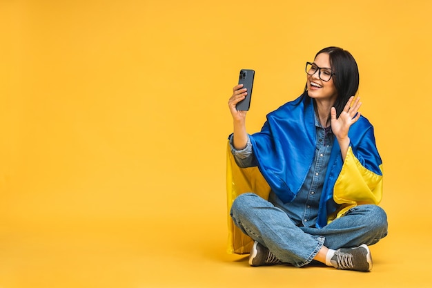 Portret van mooie mooie mooie blij vrolijke vrouw in handen Oekraïense vlag met plezier geïsoleerd op gele achtergrond Zittend op de vloer in lotus houding met behulp van mobiele telefoon