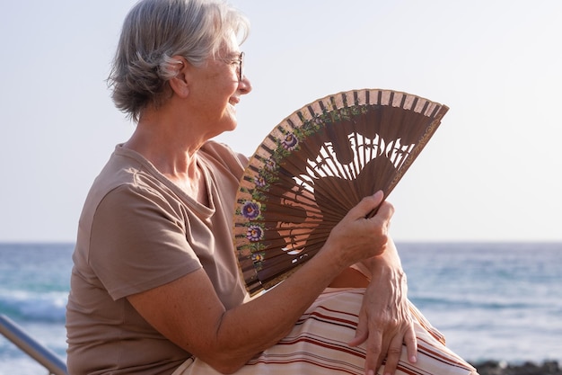 Portret van mooie oudere vrouw zittend op het strand bij zonsondergang terwijl ze lucht blaast met een ventilator, ontspannen oudere dame geniet van vakantie en vrijheid