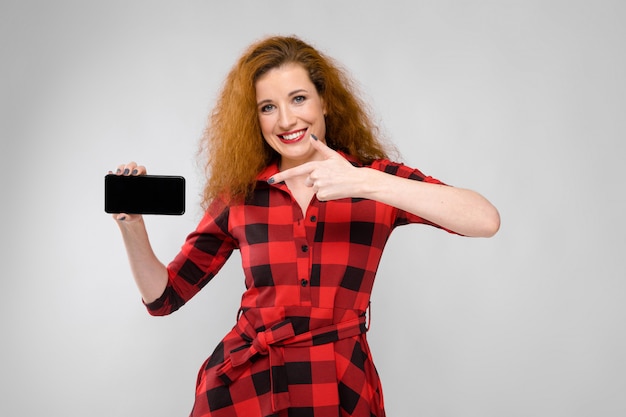 Foto portret van mooie roodharige gelukkige jonge vrouw in geruite kleding die tonend mobiele telefoon op grijze achtergrond glimlachen