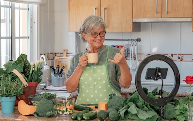 Portret van mooie senior vrouw die voor een telefoon spreekt om online inhoud van kookconcept van modern thuiswerk en digitale maker dame met glas te spreken