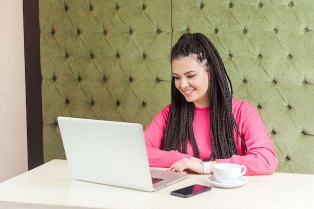 Portret van mooie tevreden jonge vrouw freelancer met zwarte dreadlocks kapsel in roze blouse zitten in café en werken op laptop met brede glimlach en kijken naar het scherm. Binnen.