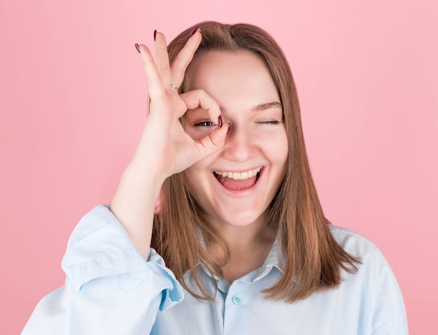 Portret van mooie vrolijke vrouw die pret heeft en door vingergaten op roze in studio kijkt