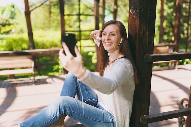 Portret van mooie vrouw die lichte vrijetijdskleding draagt. Lachende vrouw zittend in stadspark in straat buiten op lente natuur, selfie schot op mobiele telefoon of video-oproep doen. Levensstijlconcept.