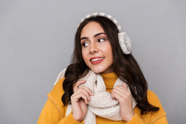 Portret van mooie vrouw die oorwarmers en sjaal draagt ?? die en sjaal glimlachen aan te raken, die over grijze achtergrond wordt geïsoleerd