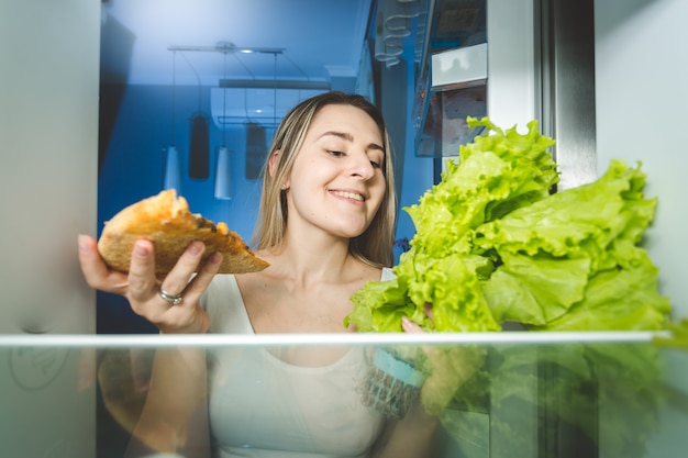 Portret van mooie vrouw die tussen pizza en verse salade kiest. Uitzicht vanaf de binnenkant van de koelkast. Concept van gezond en ongezond voedsel.