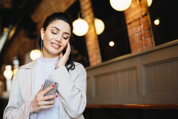 Portret van mooie vrouw met donker haar vastgebonden in paardenstaart gekleed formeel luisteren naar muziek met oortelefoons die haar ogen sluiten met ontspanning met mobiele telefoon Mensen levensstijl technologie