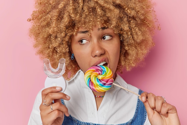 Portret van mooie vrouw met krullend haar bijt veelkleurige lolly houdt mond expander geeft niet om tanden eet schadelijk voedsel gefocust weg draagt wit overhemd geïsoleerd over roze achtergrond