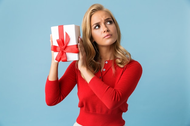 Portret van nieuwsgierige blonde vrouwen20s die rood overhemd dragen die huidige doos houden, die over blauwe muur wordt geïsoleerd