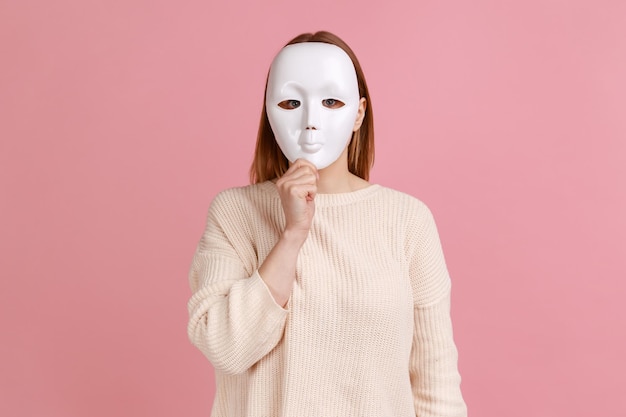 Portret van onbekende anonieme vrouw die haar gezicht bedekt met een wit masker dat haar echte persoonlijkheidsanonimiteit verbergt, gekleed in een witte trui Indoor studio-opname geïsoleerd op roze achtergrond
