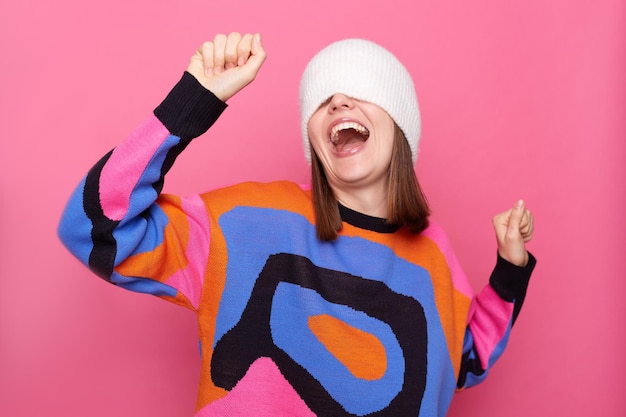 Portret van onbekende vrolijke vrouw met trui die witte pet op haar gezicht trekt en danst met opgeheven armen houdt mond open poseren geïsoleerd op roze achtergrond