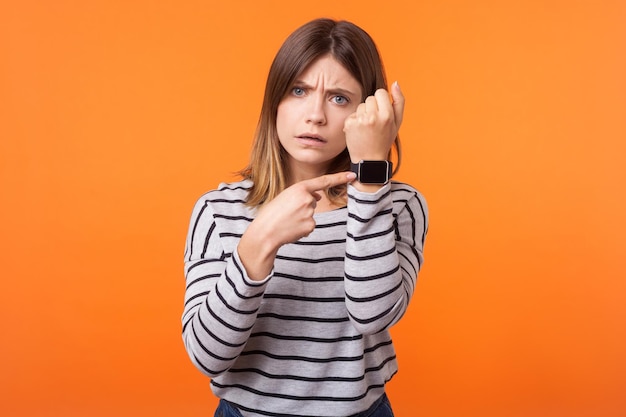 Portret van ongeduldige jonge vrouw met bruin haar in gestreept shirt met lange mouwen, wijzend op horloges om haar pols, ernstig en bezorgd binnen studio-opname geïsoleerd op oranje achtergrond