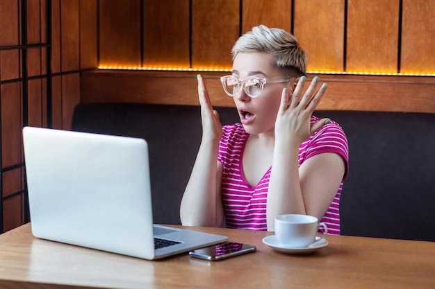 Portret van ongelooflijk jong meisje met kort haar in roze t-shirt en bril zit in café, werkt en kijkt naar video op laptop, opgeheven handen met geschokt gezicht, binnen, levensstijl