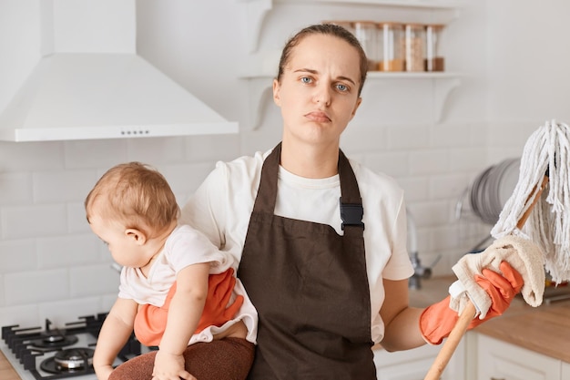 Portret van ongelukkige huisvrouw moeder met wit t-shirt en bruin schort met dweil en baby baby in handen poseren in keuken camera kijken met verstoorde uitdrukking