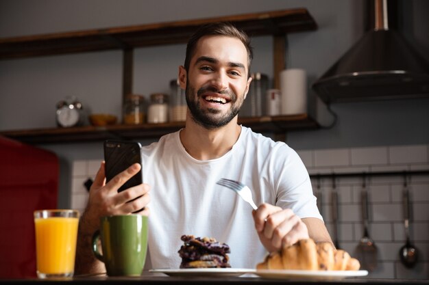 Portret van ongeschoren man 30s houden en gebruiken van smartphone tijdens het ontbijt in stijlvolle keuken thuis