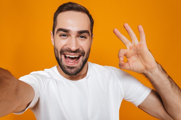 Portret van ongeschoren man in t-shirt glimlachen tijdens het nemen van selfie foto, geïsoleerd op geel