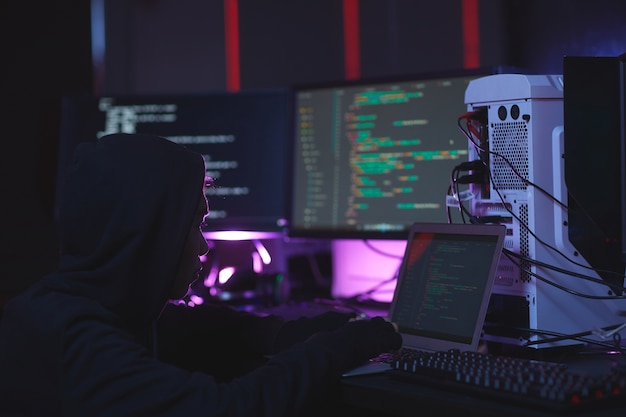 Portret van onherkenbare hacker met behulp van computerapparatuur met programmacode op schermen in de donkere kamer, cyberveiligheidsconcept, kopie ruimte