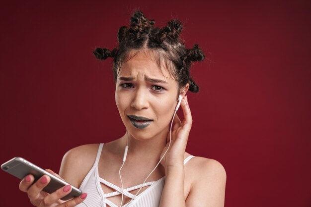 Portret van ontevreden punkmeisje met bizar kapsel en donkere lippenstift die fronst tijdens het gebruik van smartphone met koptelefoon geïsoleerd over rode muur