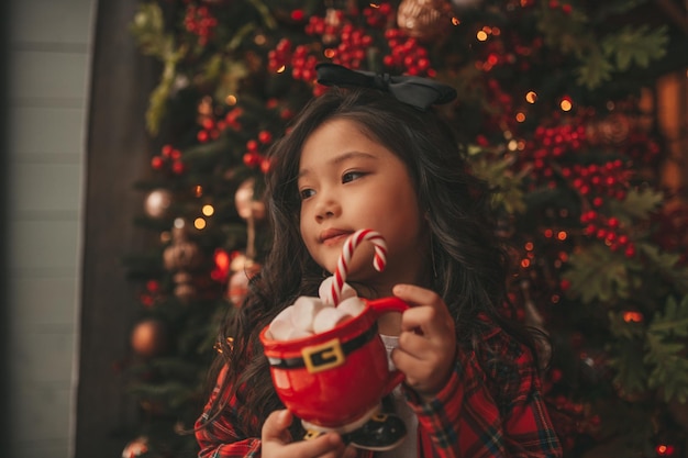 Portret van openhartig aziatisch glimlachend meisje houdt rode kerstmok vast met marshmallows en snoepgoed