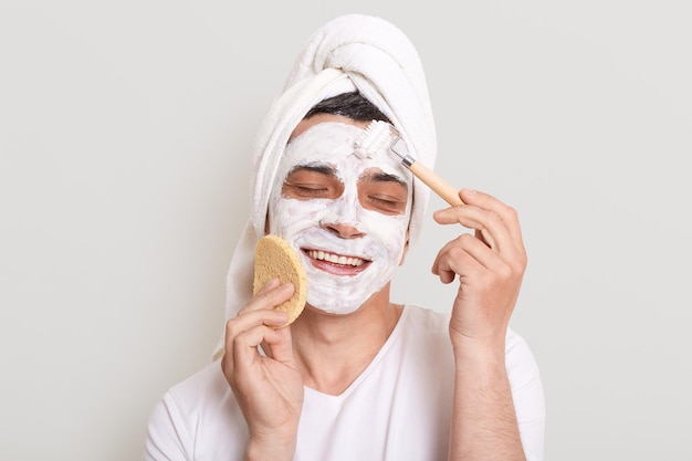 Portret van opgetogen tevreden man met wit t-shirt poseren geïsoleerd over grijze achtergrond permanent met masker op gezicht procedures doen voor huidverzorging met behulp van spons en massage roller genieten