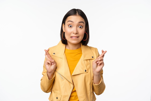 Portret van opgewonden aziatische vrouw ziet er hoopvol uit terwijl ze bidt of smeekt, wachtend op nieuws dat op een witte achtergrond staat en enthousiast glimlacht