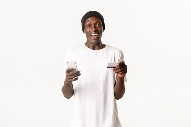 Portret van opgewonden, gelukkige Afro-Amerikaanse jonge mannelijke student, met creditcard en smartphone met geamuseerde glimlach