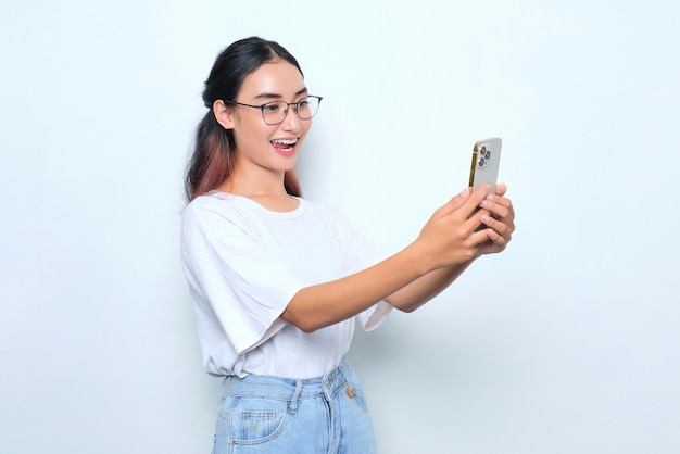 Portret van opgewonden jong Aziatisch meisje in witte t-shirt met behulp van mobiele telefoon geïsoleerd op een witte achtergrond