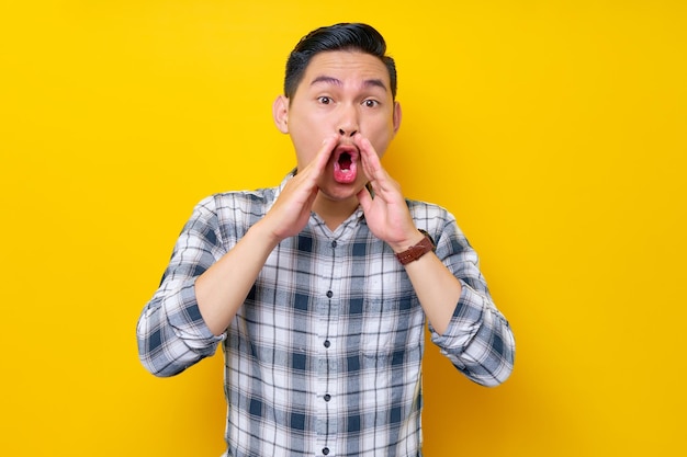 Foto portret van opgewonden jonge aziatische man in geruite overhemd schreeuwend nieuws over verkoopkortingen met handen in de buurt van mond geïsoleerd op gele achtergrond mensen levensstijl concept