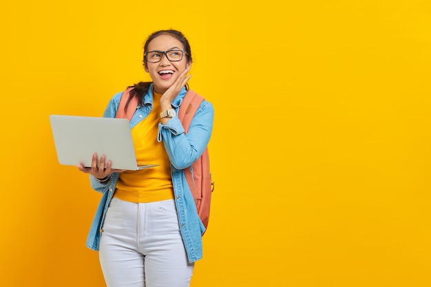 Portret van opgewonden jonge Aziatische vrouw student in casual kleding met rugzak met behulp van laptop en wang aan te raken met handen geïsoleerd op gele achtergrond Onderwijs in college universiteit concept