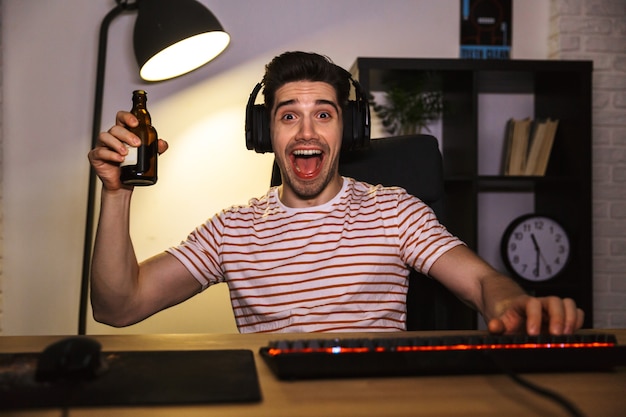 Portret van opgewonden jonge kerel die hoofdtelefoon draagt die bier drinkt, terwijl hij aan bureau met computer in kamer zit en camera bekijkt