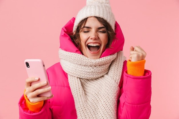 Portret van opgewonden jonge vrouw in warme kleren die schreeuwt en mobiele telefoon gebruikt die op roze wordt geïsoleerd