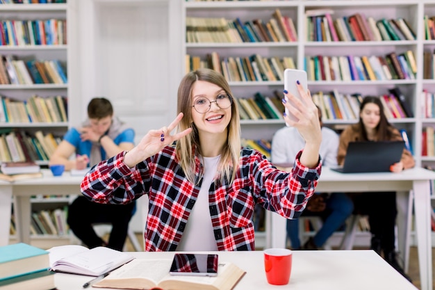 Portret van opgewonden lachende jonge mooie blonde vrouw in geruit hemd en bril selfie foto maken en v-teken met twee vingers tonen, zittend aan de tafel in de bibliotheek leeszaal