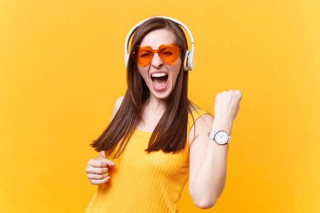 Portret van opgewonden leuk meisje in oranje bril die muziek luistert in koptelefoon die winnaargebaar doet, zeg ja, kopieer ruimte geïsoleerd op gele achtergrond. Mensen oprecht emoties concept. Reclame gebied.