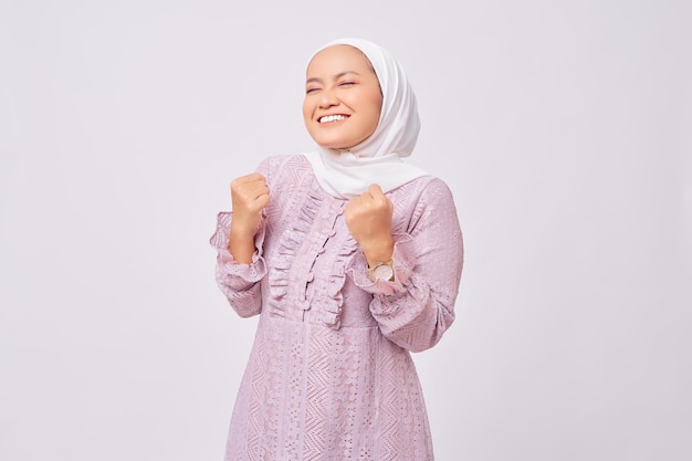 Portret van opgewonden mooie jonge Aziatische moslimvrouw die hijab en paarse jurk draagt, vuisten opsteekt en de overwinning viert op Ramadan geïsoleerd op witte studio achtergrond