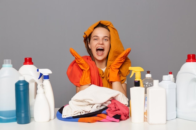 Portret van opgewonden verbaasde grappige vrouw zittend op de werkplek met vuile kleding op haar hoofd gebruikt verschillende wasmiddelen voor het doen van huishoudelijke klusjes geïsoleerd over grijze achtergrond