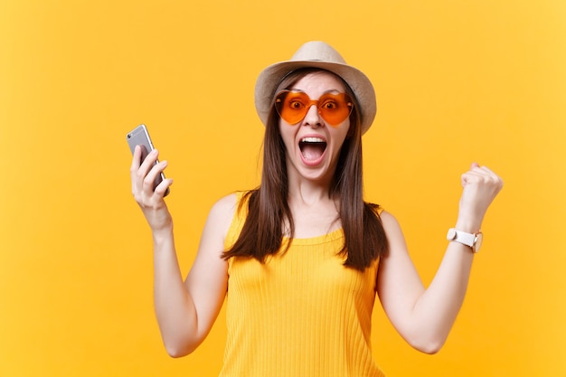 Portret van opgewonden verbaasde jonge vrouw in stro zomerhoed, oranje bril die mond wijd open houdt, verbaasd kijkt, mobiele telefoon geïsoleerd op gele achtergrond houdt. Mensen oprecht emoties concept.