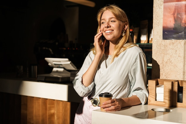 Portret van optimistische jonge vrouw die lacht en praat op smartphone terwijl ze afhaalkoffie drinkt in café