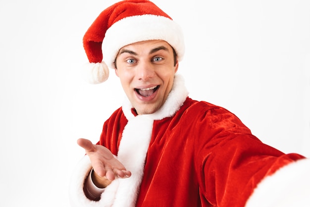 Portret van optimistische man 30s in kerstman kostuum en rode hoed lachen tijdens het nemen van selfie foto