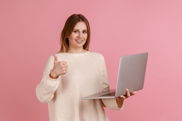 Portret van optimistische mooie jonge volwassen blonde vrouw die op laptop compute werkt en naar de camera kijkt en duim toont die een witte trui draagt. Indoor studio shot geïsoleerd op roze achtergrond