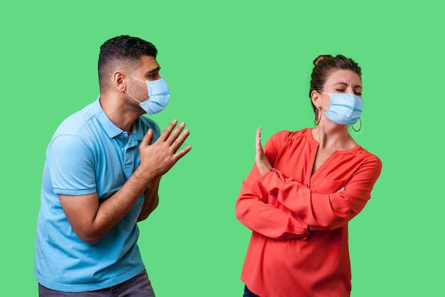 Portret van paar met chirurgisch medisch masker dat samen staat, man met armen in gebed en smekend om vergeving, kijkend met hoop naar ondeugende vrouw, geïsoleerd op groene achtergrond, indoor schot