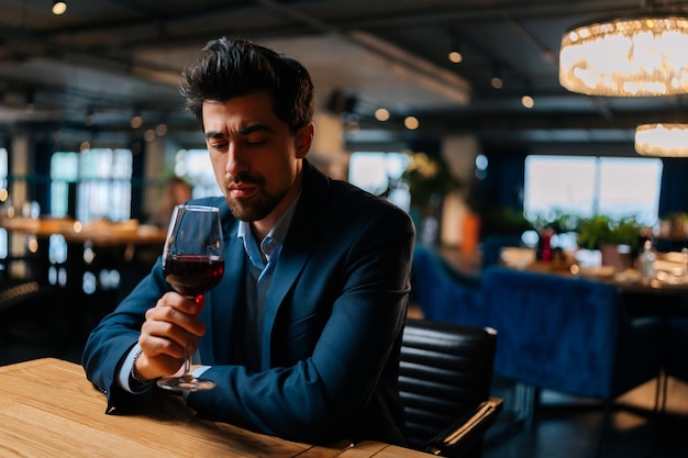 Portret van peinzende elegante man in pak met in de hand glazen rode wijn zittend aan tafel in restaurant met donker interieur bedachtzaam naar beneden kijkend