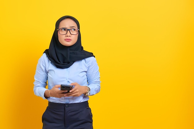 Portret van peinzende jonge Aziatische vrouw die een mobiele telefoon gebruikt en omhoog kijkt geïsoleerd op gele achtergrond