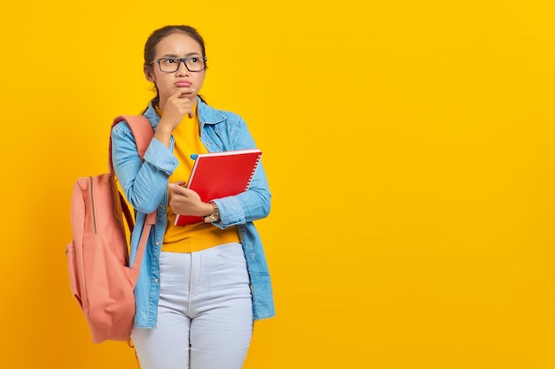 Portret van peinzende jonge Aziatische vrouw student in casual kleding met rugzak boek te houden en na te denken over vraag geïsoleerd op gele achtergrond Onderwijs in college universiteit concept