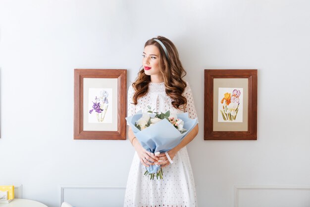 Portret van peinzende jonge vrouw in shite-jurk die staat en een boeket bloemen vasthoudt