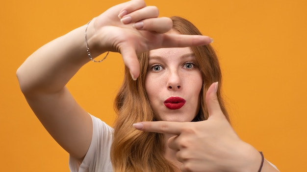 Portret van positief meisje met golvende roodharige, wit t-shirt dragen, heeft een goed humeur, gebaren vinger frame actief op camera.