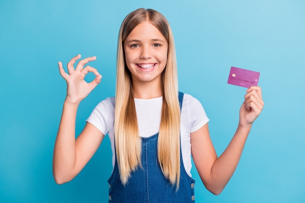 Portret van positief mooi blond haar meisje houdt bankkaart tonen ok teken kopen te koop geïsoleerd op pastel blauwe kleur achtergrond